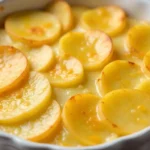 A close-up of a baked dish featuring thinly sliced potatoes seasoned with cheese powder and spices, arranged in a white baking dish.